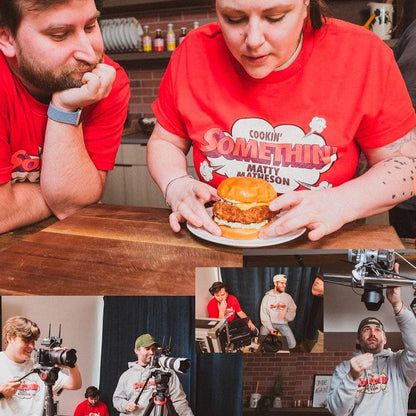 Two people in red Cookin Somethin Original T-shirts enjoying a burger at a wooden table, with additional photos showing behind-the-scenes filming moments of the YouTube cooking show. Product is displayed in both casual dining and production settings.