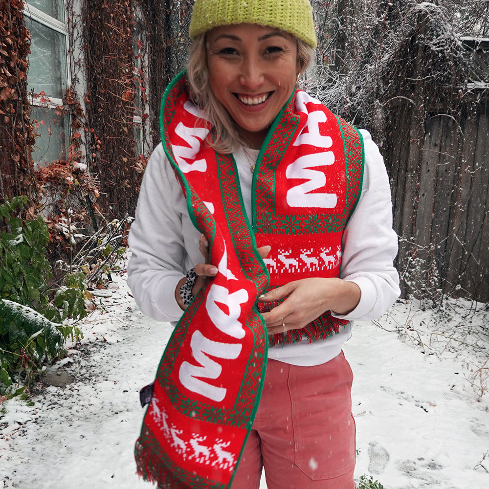 A smiling person stands outside in the snow, wearing an exclusiv Matty Matheson merch with a holiday pattern.. Surrounded by snow, their vibrant winter look is highlighted by the Matty Holiday Scarf from Mattys World, adding festive red and green to the scene.