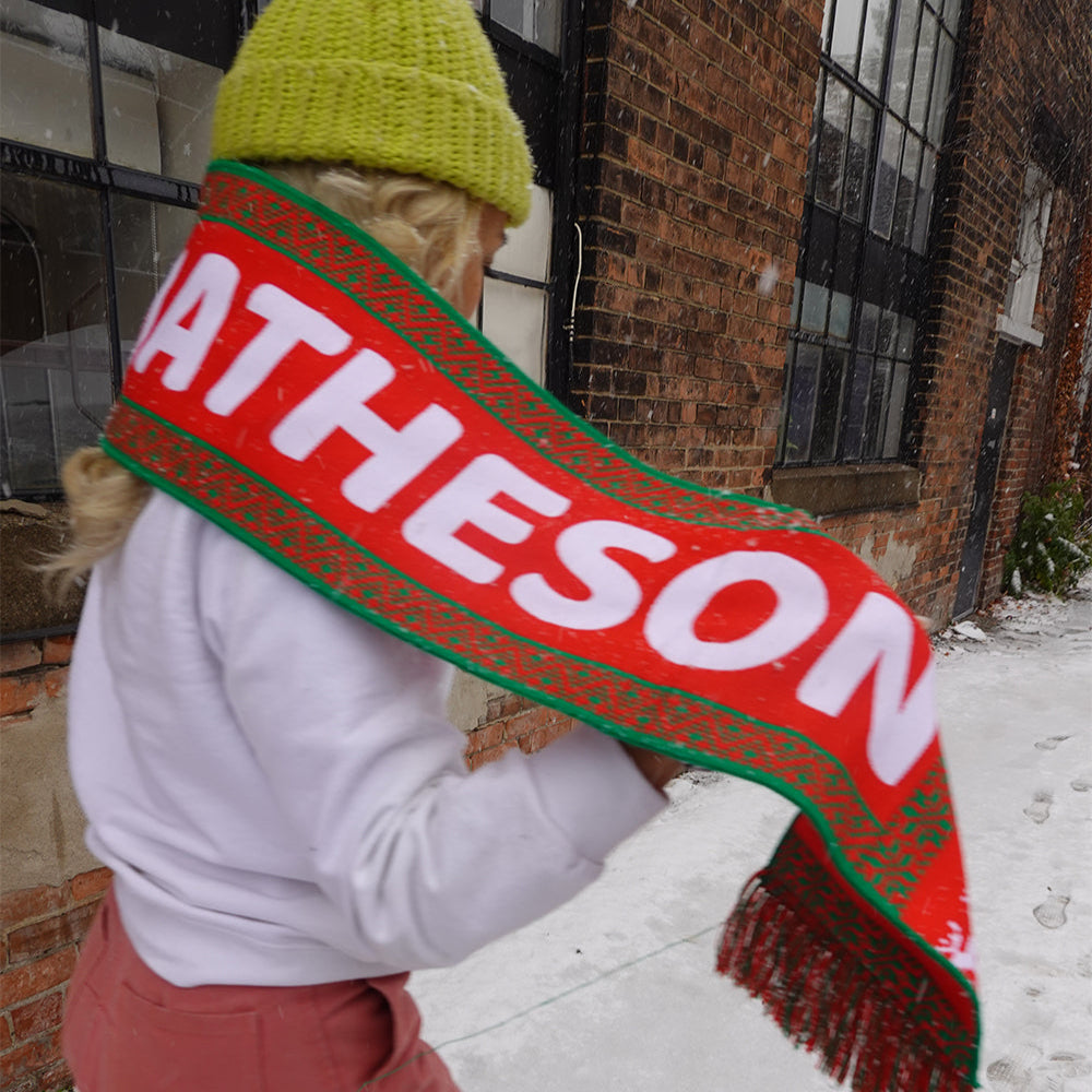 A person strolling on a snowy path holding Mattys Worlds Matty Holiday Scarf. This 100% acrylic scarf with white letters adds a splash of color perfect holiday party wear from Matty Mathesons exclusive merch collection.
